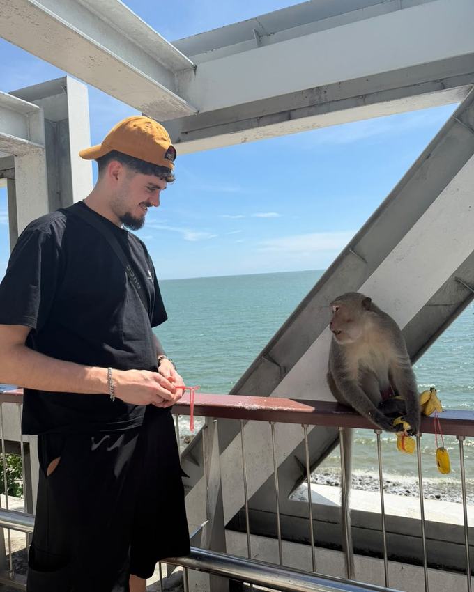Steve Kotzorek, in a cap, on a seaside platform in Thailand, next to a macaque holding a banana